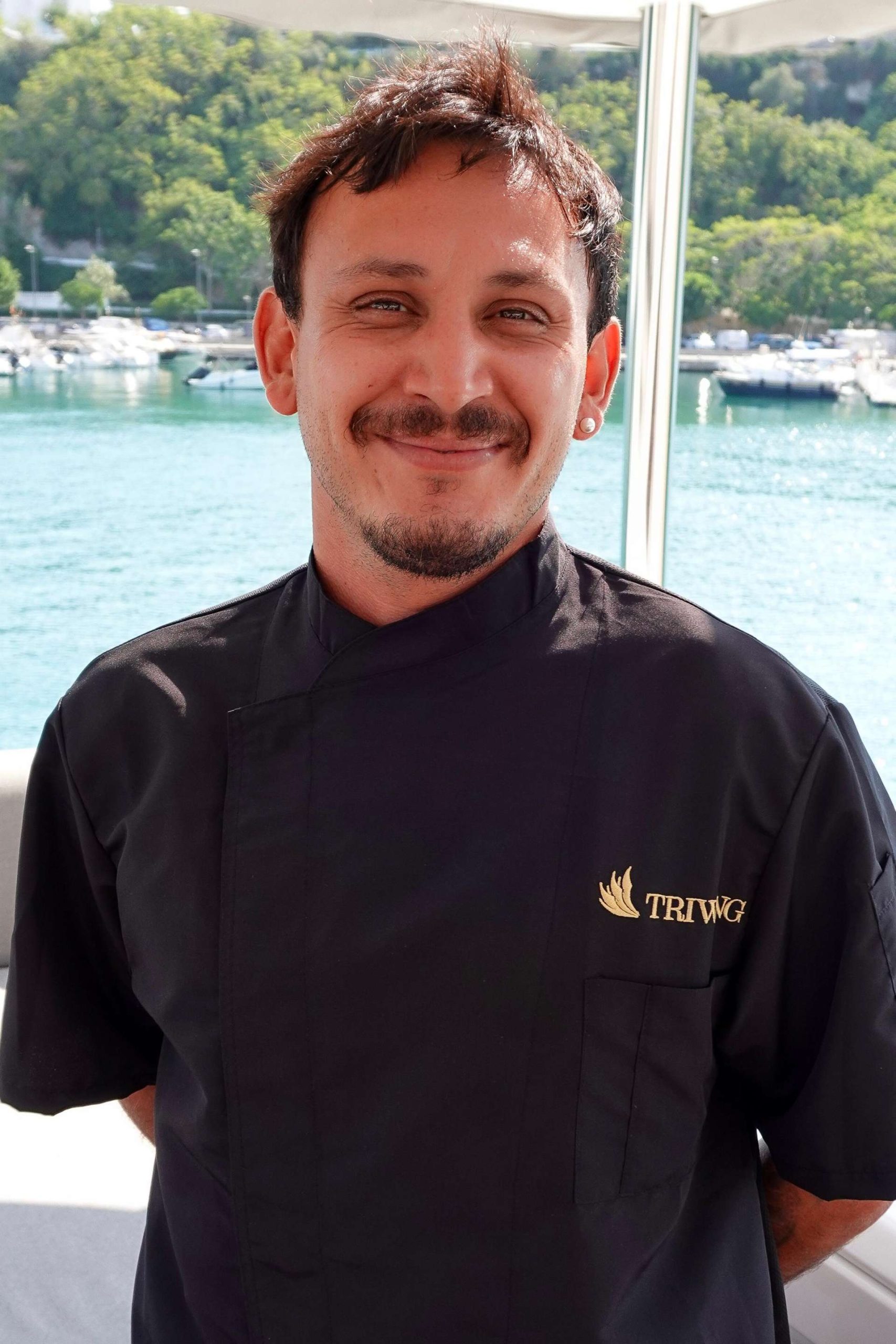Professional chef aboard the luxury catamaran Triwing Endor, wearing chef uniform with marina and turquoise water in the background during a private Caribbean yacht charter.