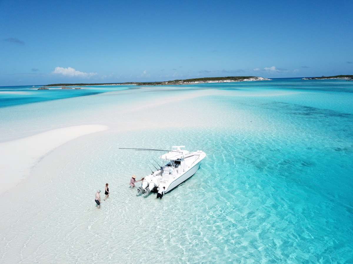 Guests exploring a shallow turquoise sandbar beside a small boat in the Bahamas during a luxury yacht charter adventure in crystal-clear Caribbean waters.